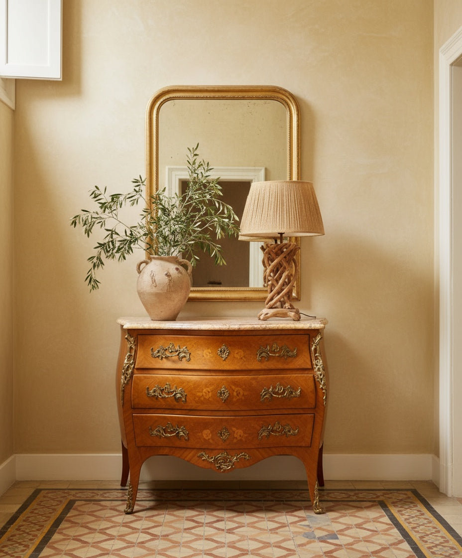 ómoda Luis XV bombé en un recibidor clásico con espejo dorado, lámpara de madera tallada y jarrón rústico.