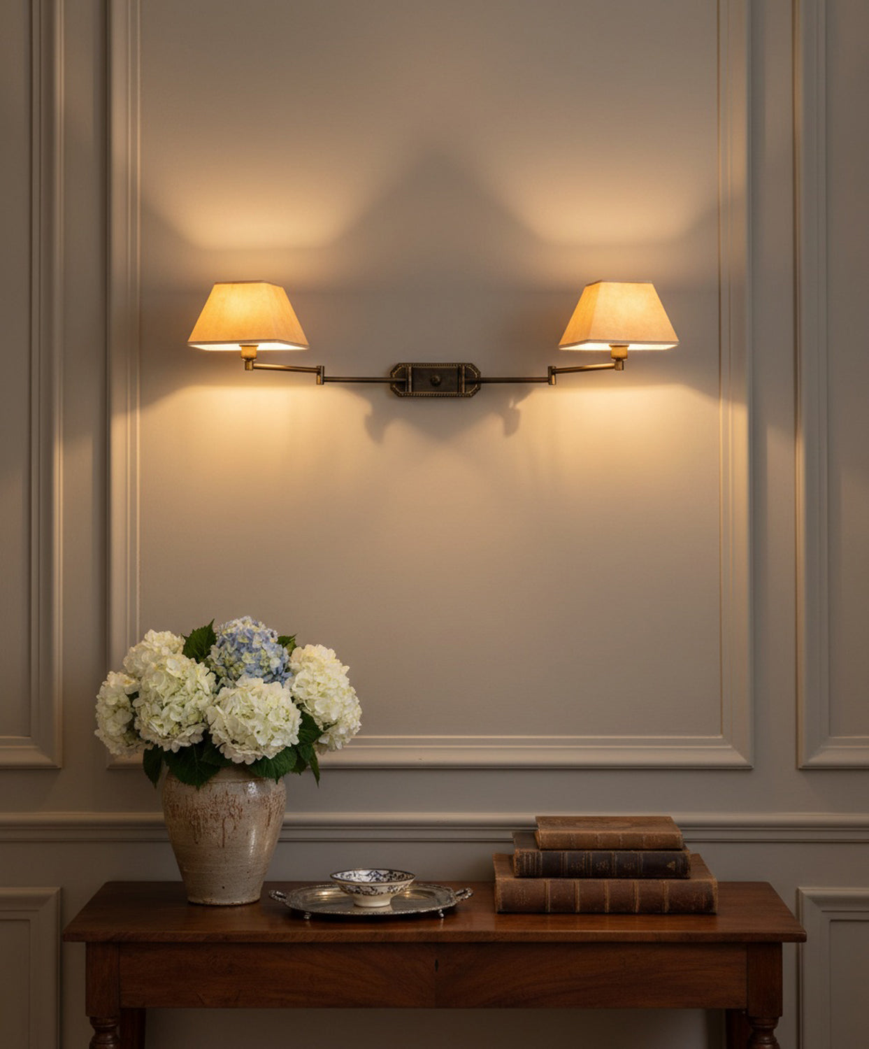 Lámpara de pared doble encendida sobre consola de madera clásica con libros antiguos y jarrón de hortensias.