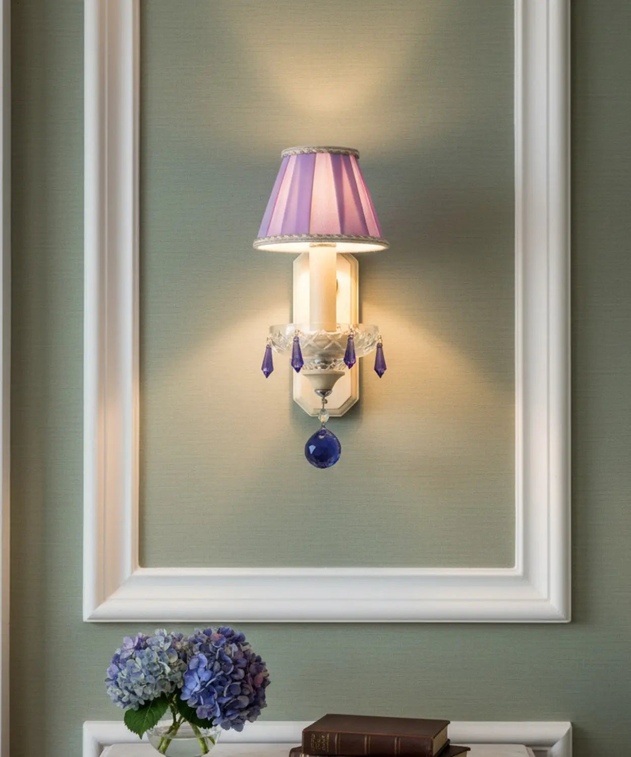 Aplique iluminado instalado sobre una pared verde musgo enmarcado en molduras blancas, flanqueado por hortensias azules.