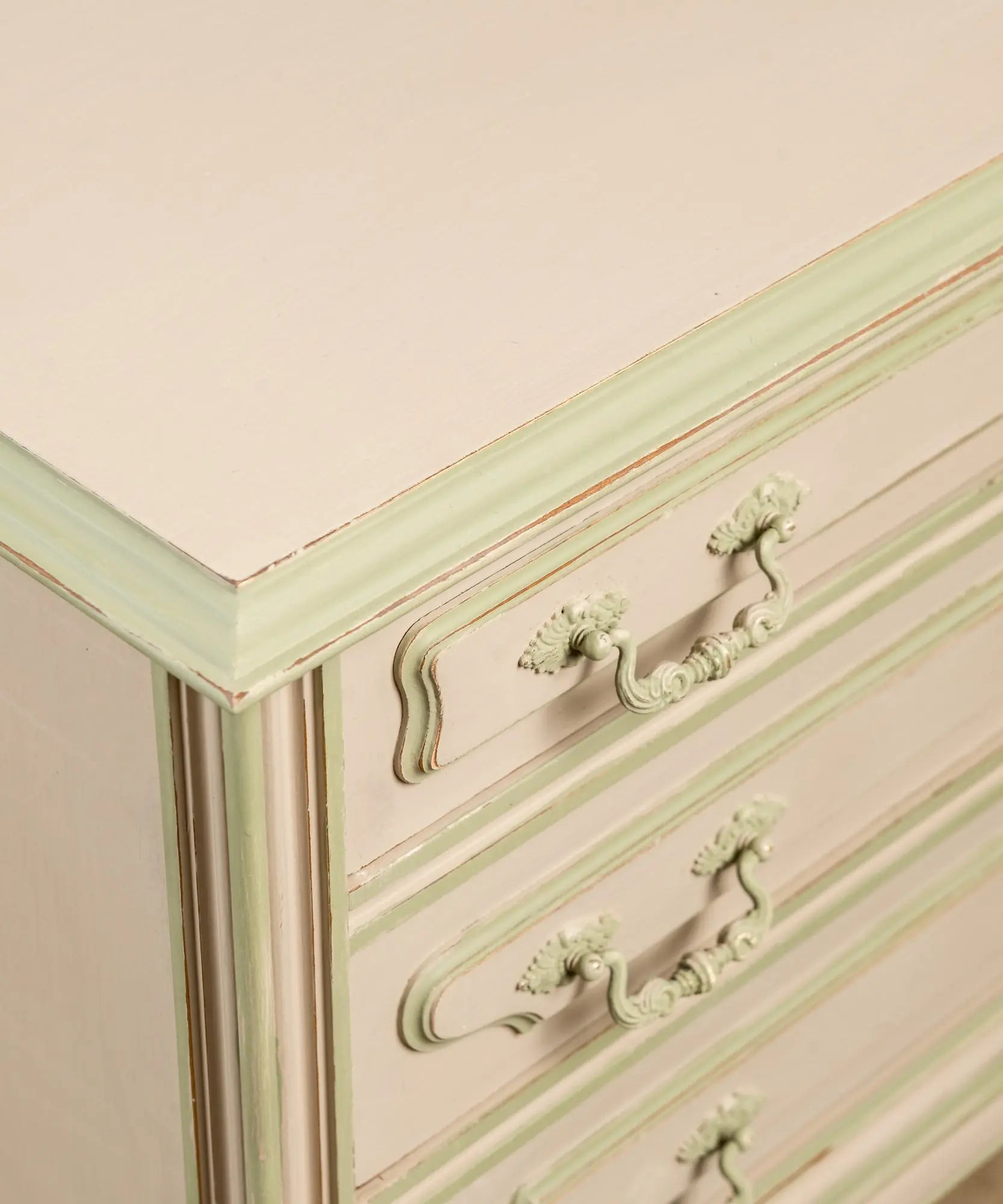 Primer plano de tiradores clásicos y molduras pintadas en verde sobre cajones color beige arena.