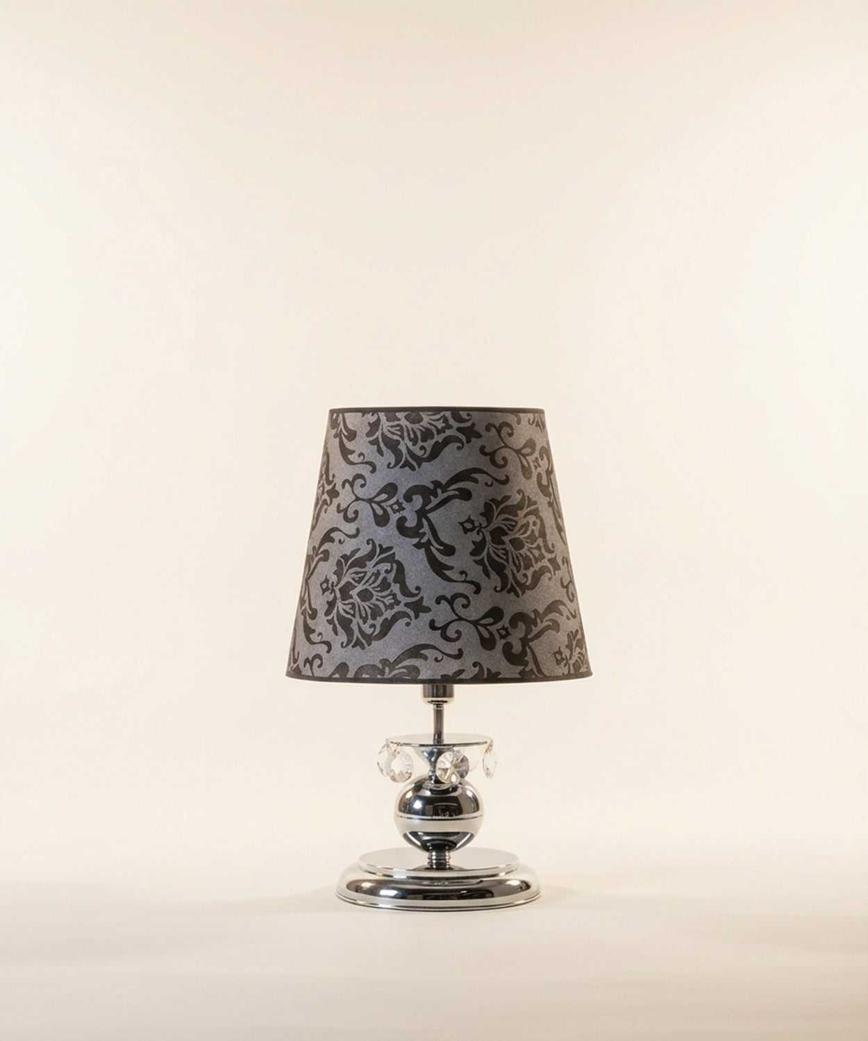Lámpara de sobremesa de diseño neobarroco con base cromada esférica, detalles de cristales de Swarovsky colgantes y pantalla gris con estampado damasco negro, fondo neutro.