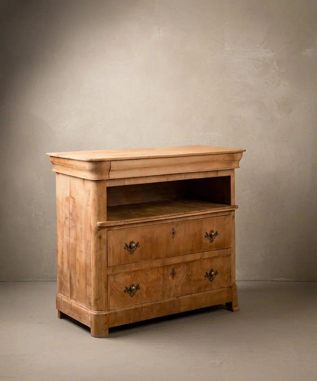 Cómoda antigua de madera clara mostrando el cajón superior oculto en gola y herrajes de bronce.