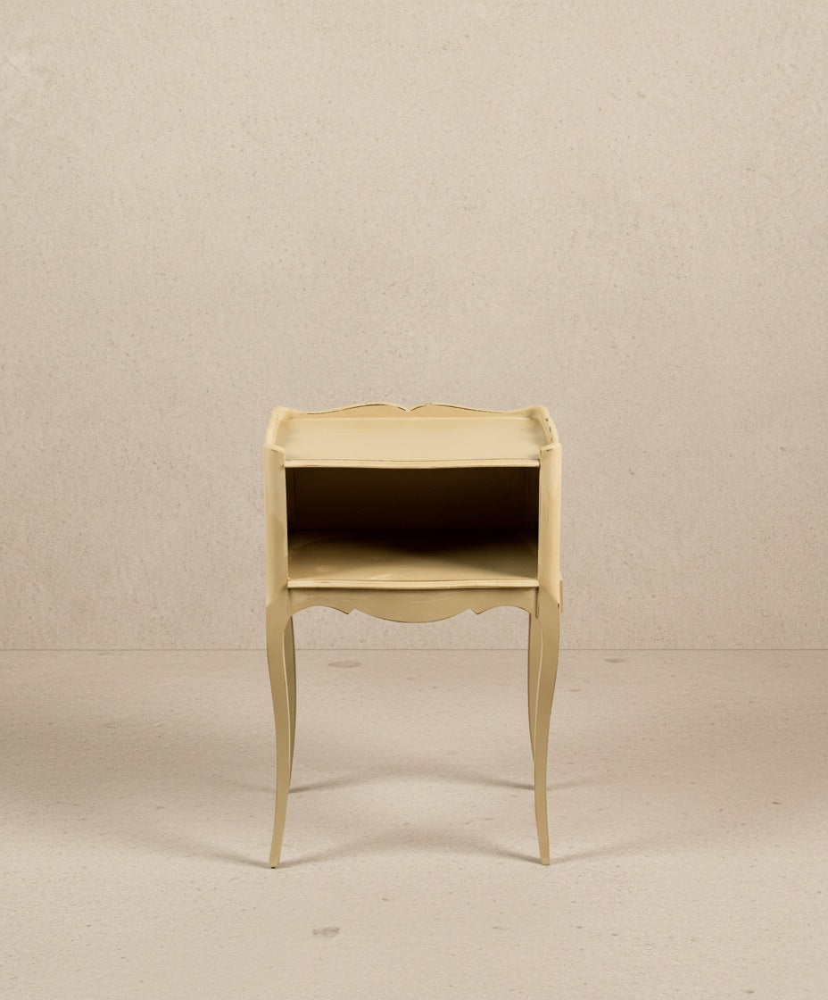 Vista Frontal: Mesilla de noche color beige con bordes festoneados y estructura esbelta francesa.