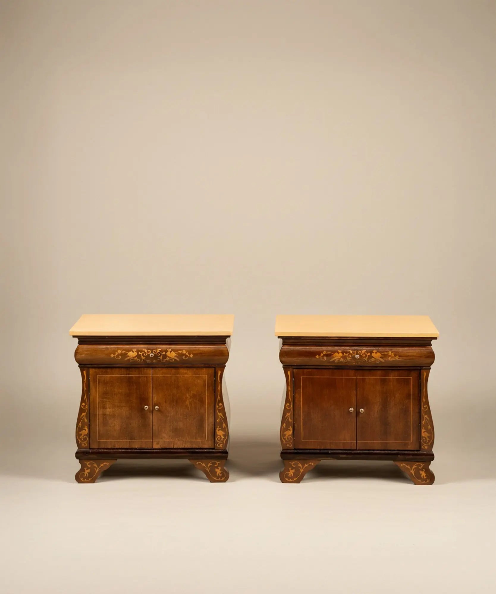 Pareja de mesitas de noche vintage con marquetería de aves y puertas abiertas frente a mural sepia.