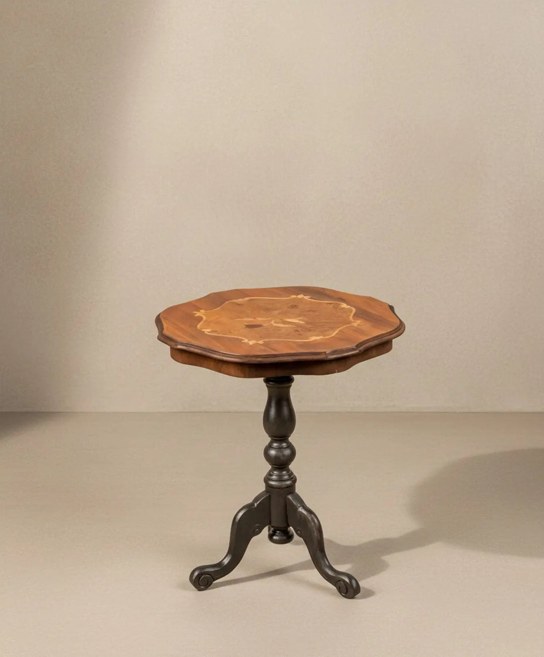 Mesa velador antigua con tablero de marquetería floral y base trípode negra sobre fondo neutro.