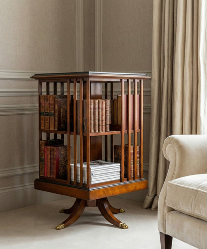 Mesa giratoria con libros antiguos junto a un sillón verde esmeralda en un salón elegante con molduras.