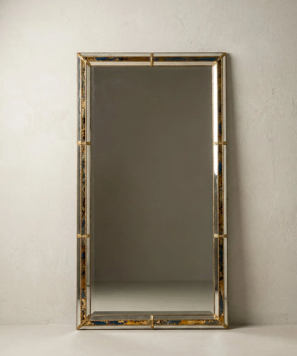 Espejo gigante rectangular apoyado en la pared en formato vertical con esquinas de latón dorado.