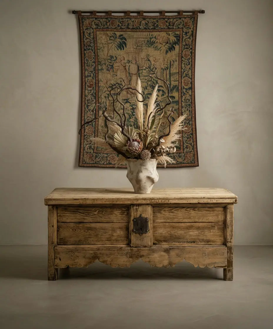 Arcón antiguo utilizado como consola bajo tapiz histórico, decorado con jarrón artesanal y flores secas.