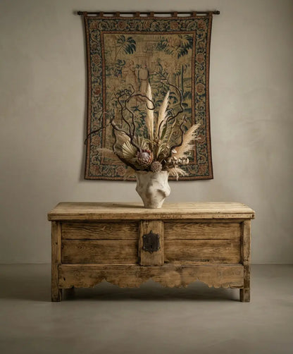 Arcón antiguo utilizado como consola bajo tapiz histórico, decorado con jarrón artesanal y flores secas.