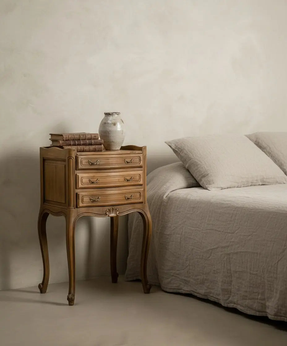 Una mesita de noche Luis XV ambientada junto a una cama con lino natural, libros antiguos y jarrón cerámico, estilo mediterráneo.