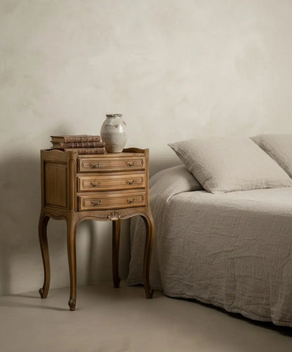 Una mesita de noche Luis XV ambientada junto a una cama con lino natural, libros antiguos y jarrón cerámico, estilo mediterráneo.