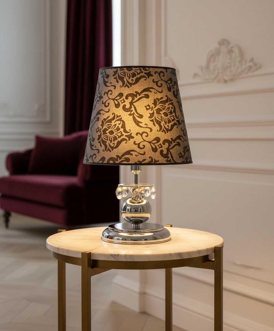 Lámpara de sobremesa de diseño neobarroco con base cromada esférica, detalles de cristales de Swarovsky colgantes y pantalla gris con estampado damasco negro sobre mesita auxiliar
