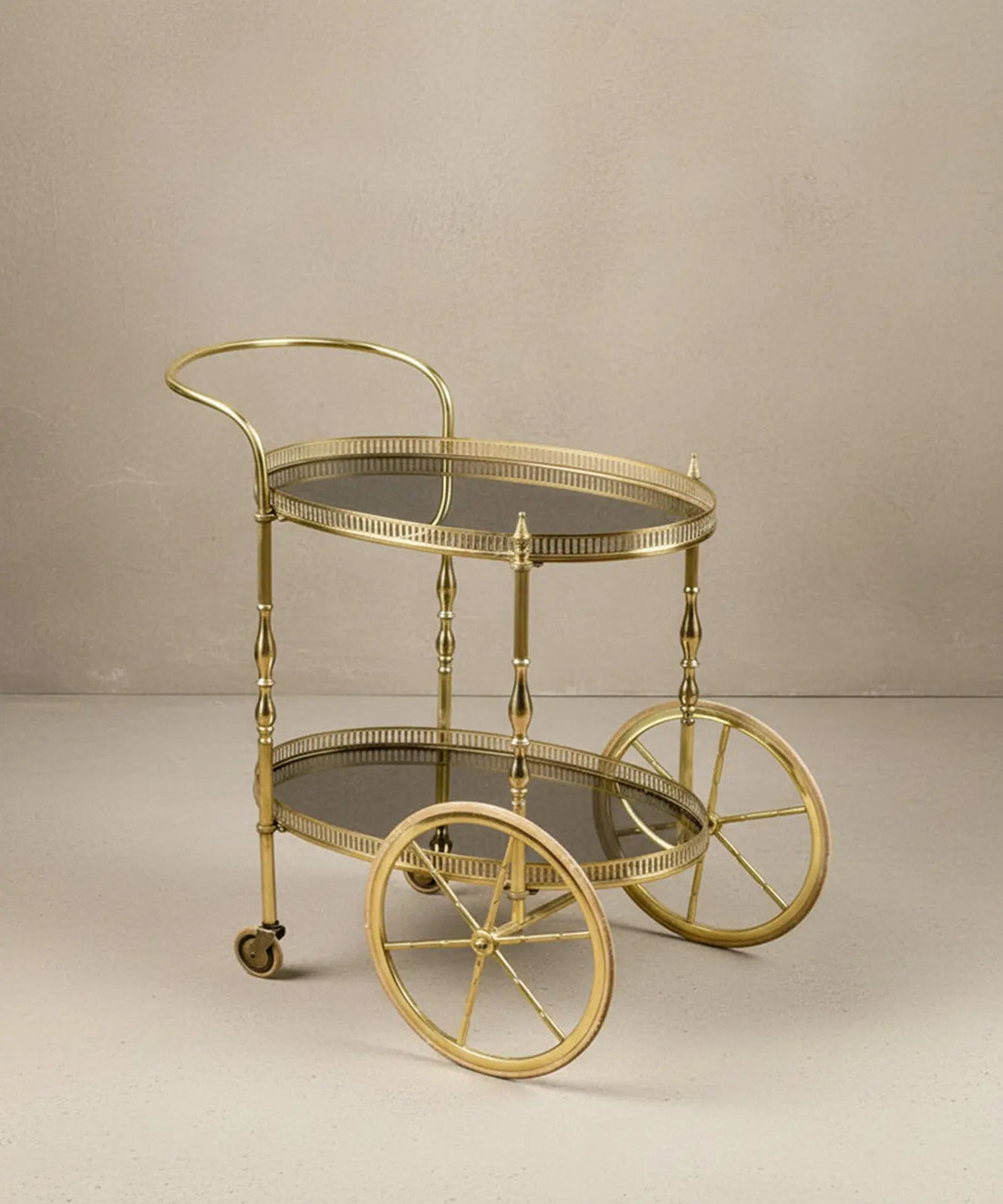 Carrito de bar vintage de latón dorado con dos niveles de cristal ahumado sobre fondo neutro.