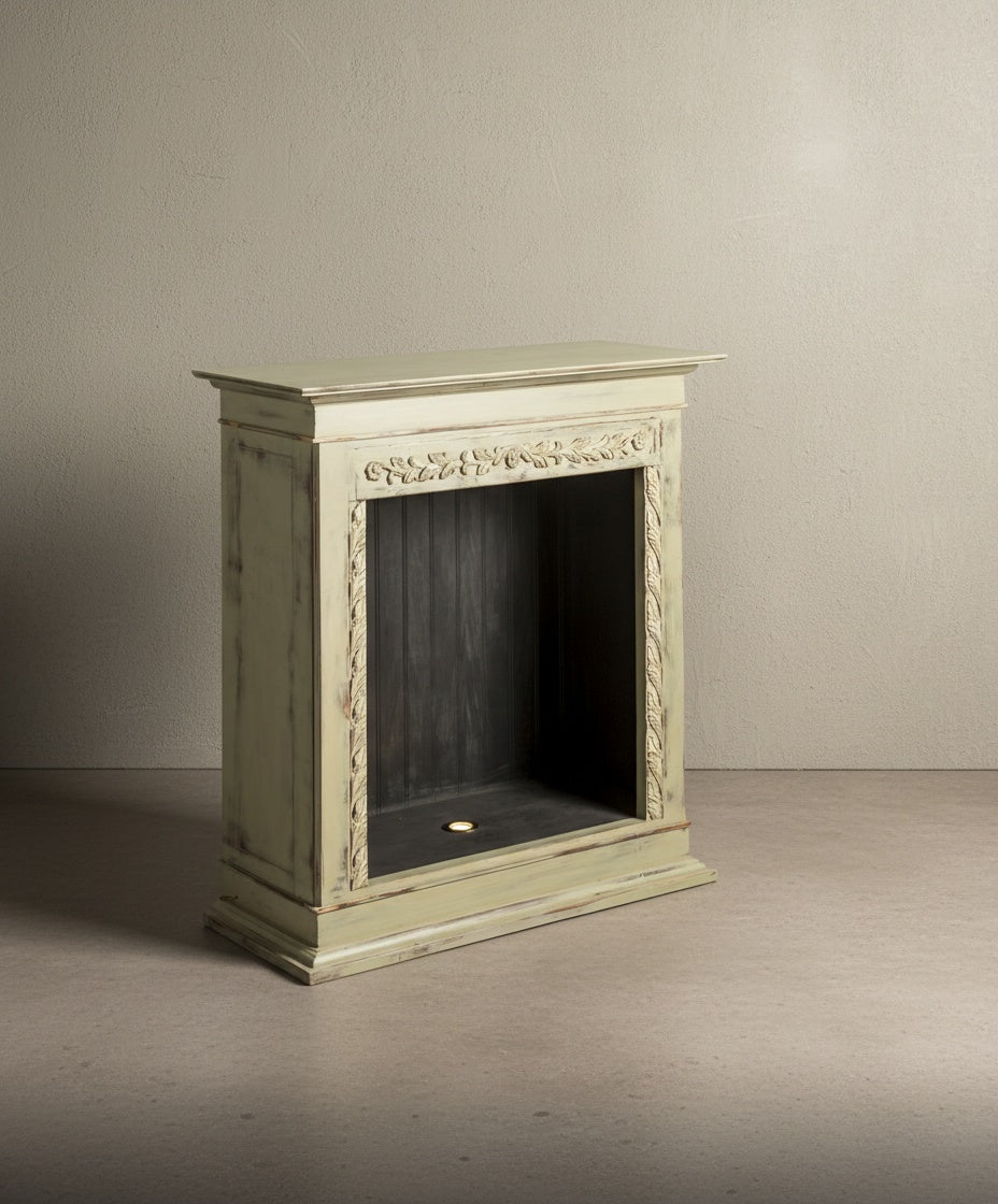  Vista lateral de la chimenea decorativa verde pátina con talla floral sobre fondo neutro.