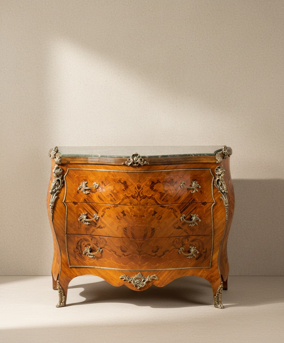 Vista frontal de cómoda bombé estilo Luis XV con marquetería de maderas nobles y tapa de mármol verde sobre fondo neutro.