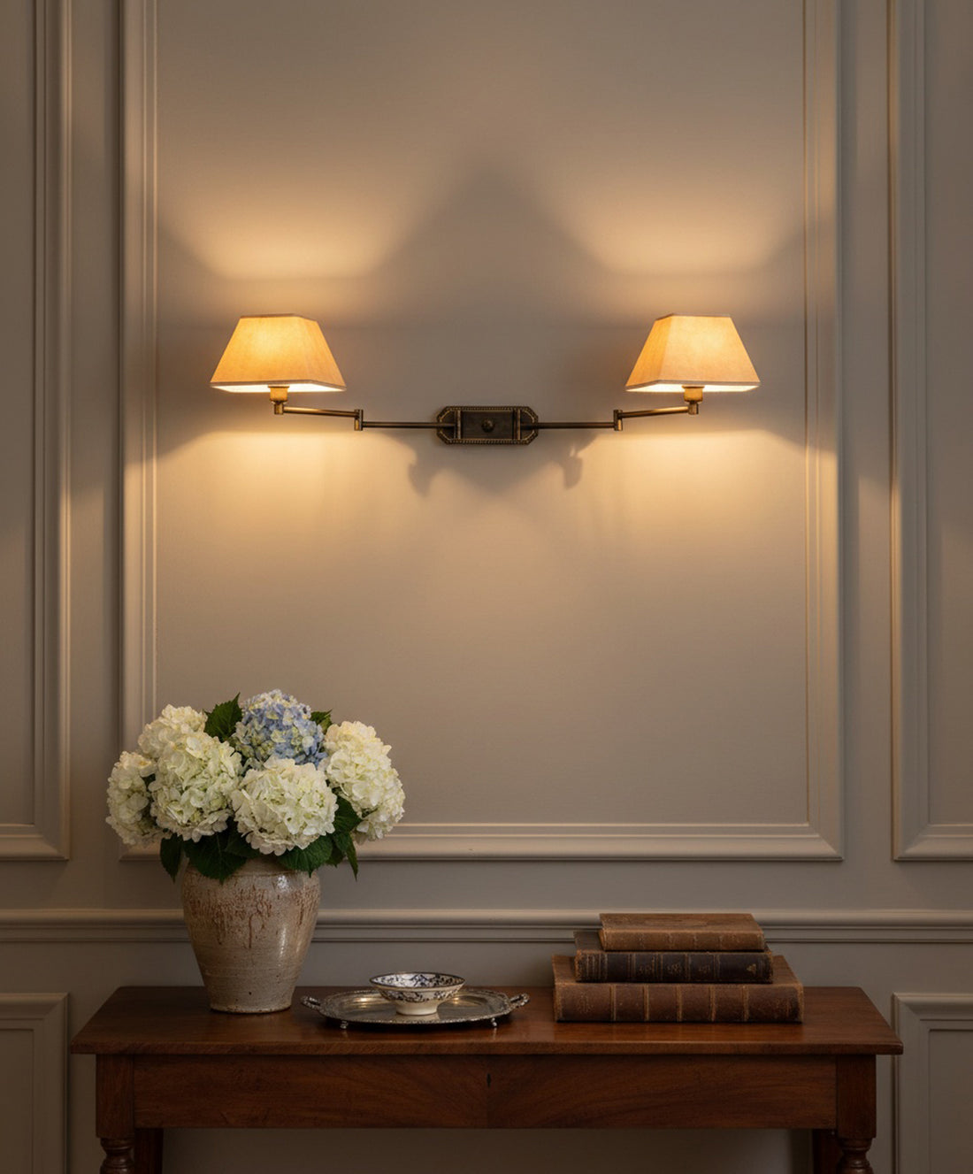 Lámpara de pared doble encendida sobre consola de madera clásica con libros antiguos y jarrón de hortensias.