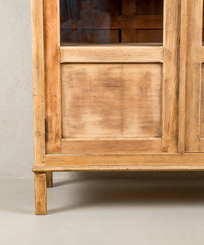 detalle Vitrina de madera clara estilo provenzal con puertas de cristal y herrajes ornamentados sobre fondo neutro.