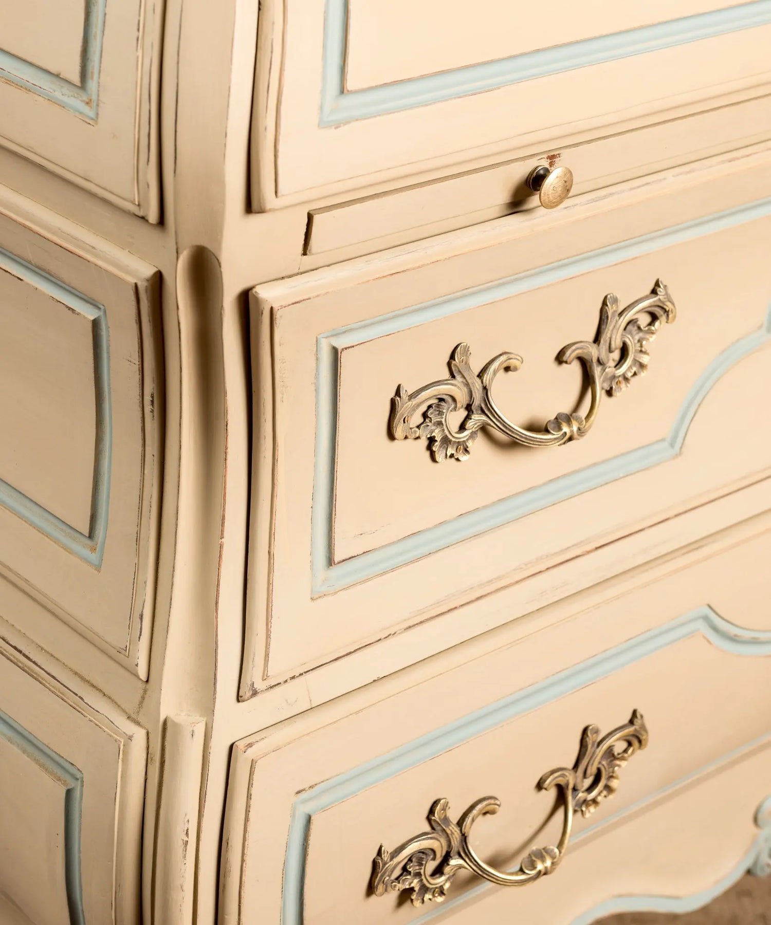 Primer plano de tirador y bocallave de bronce rococó sobre cajón de madera pintada en beige y azul decapé.