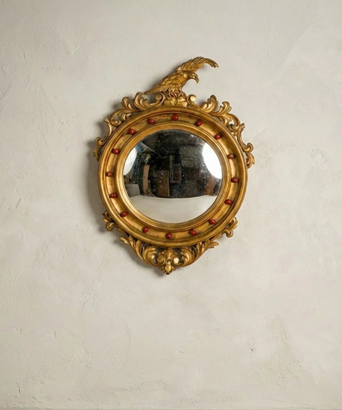 Espejo convexo ojo de bruja vintage de madera dorada coronado por un águila sobre pared clara.