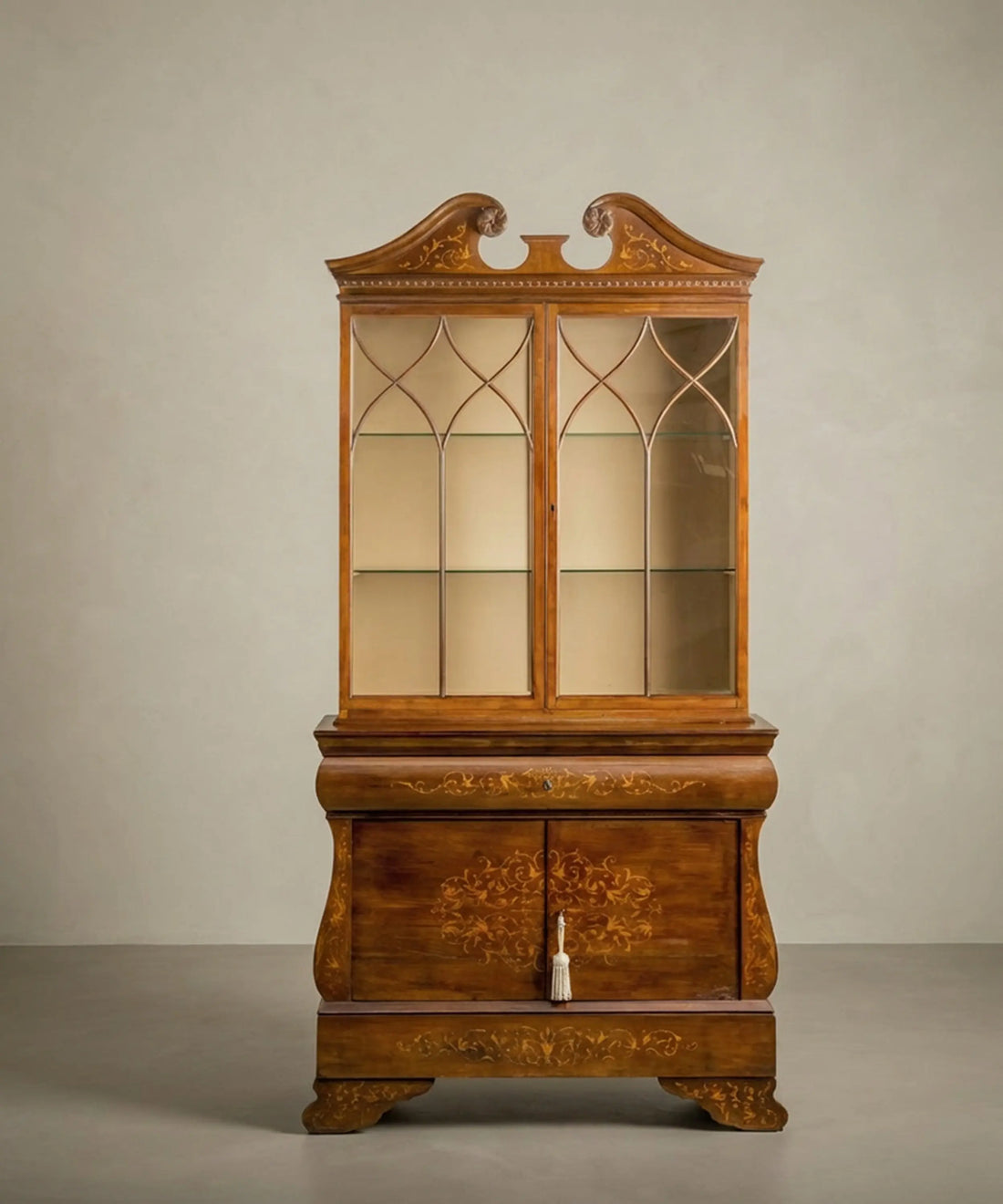 Vitrina antigua de marquetería floral estilo neoclásico con puertas acristaladas y frontón partido sobre fondo neutro.