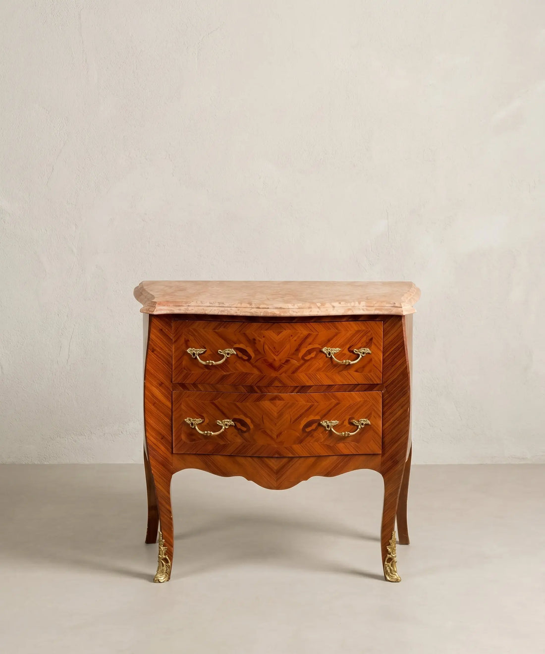 Cómoda bombé vintage de madera noble con dos cajones, bronces dorados y tapa de mármol rosado.