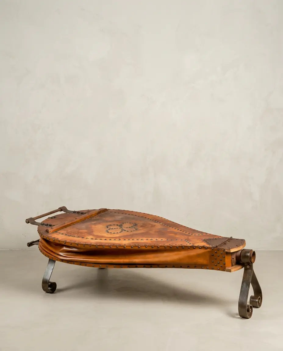 Mesa de centro fuelle de fragua antiguo en cuero marrón con patas de hierro forjado sobre fondo neutro.