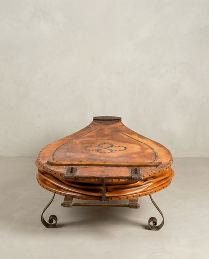 Mesa de centro fuelle de fragua antiguo en cuero marrón con patas de hierro forjado sobre fondo neutro.