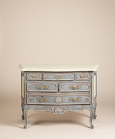 Vista frontal de una cómoda restaurada estilo Luis XV con tapa color crema y siete cajones sobre un fondo neutro.
