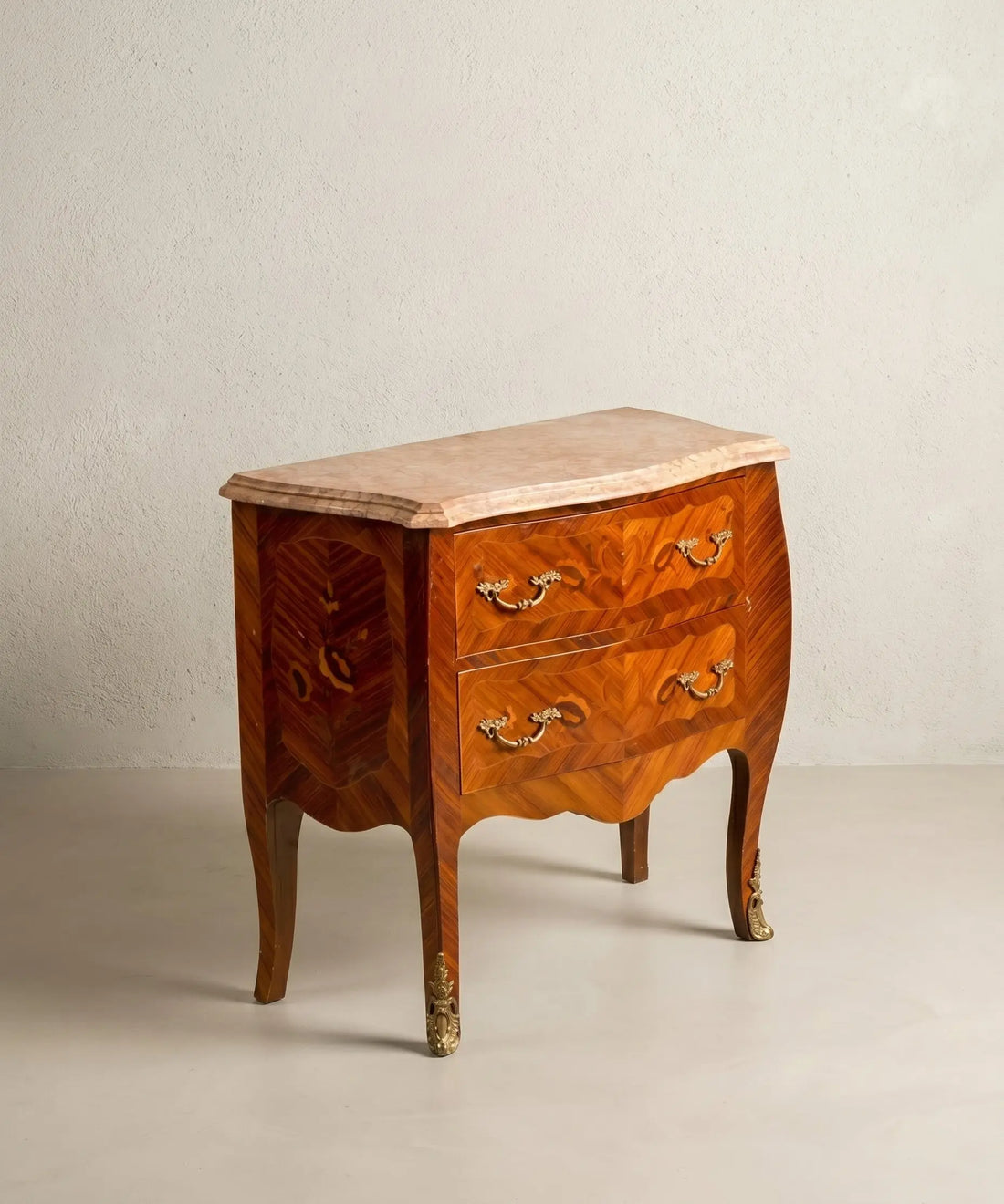 Cómoda bombé vintage de madera noble con dos cajones, bronces dorados y tapa de mármol rosado.