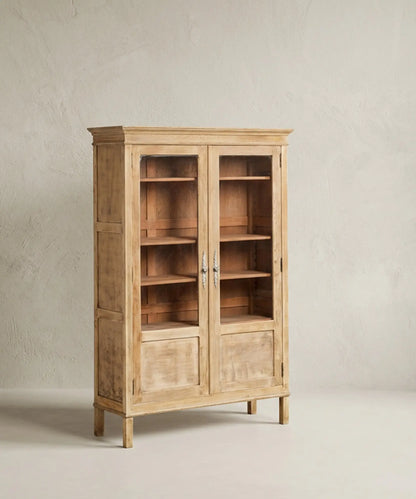 Vitrina de madera clara estilo provenzal con puertas de cristal y herrajes ornamentados sobre fondo neutro.