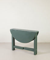 Mesa circular de alas abierta en color verde bosque con base en aspa de madera.