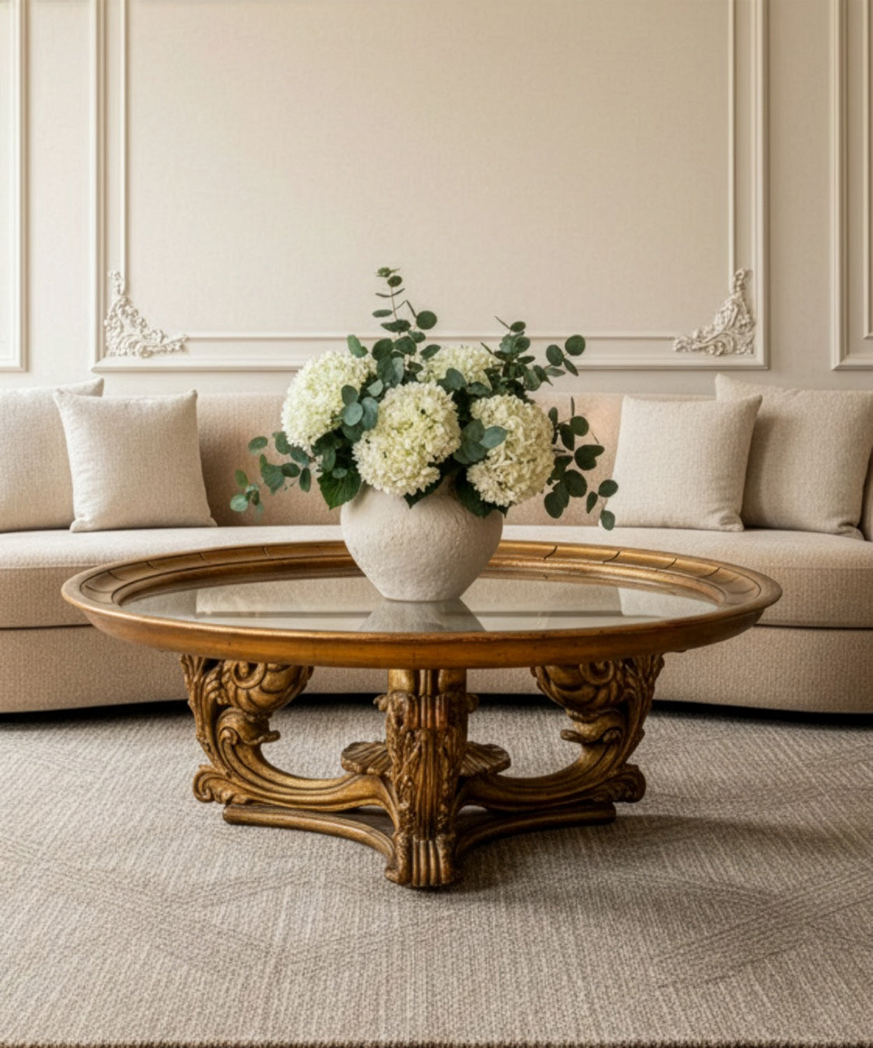 Vista frontal de mesa de centro circular dorada con base tallada y flores blancas en salón neoclásico.