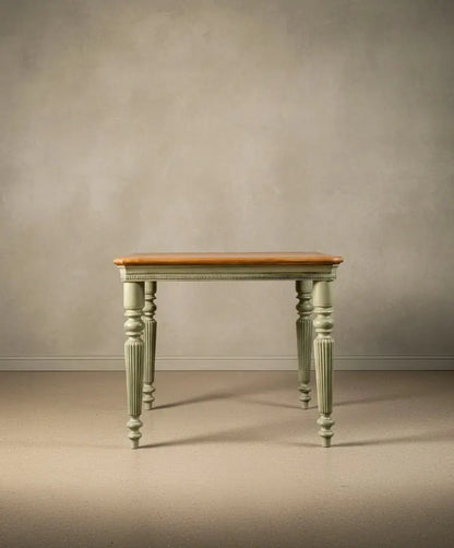 Vista frontal de mesa cuadrada de estilo provenzal con patas acanaladas en color verde musgo y tablero de madera clara natural sobre fondo neutro.