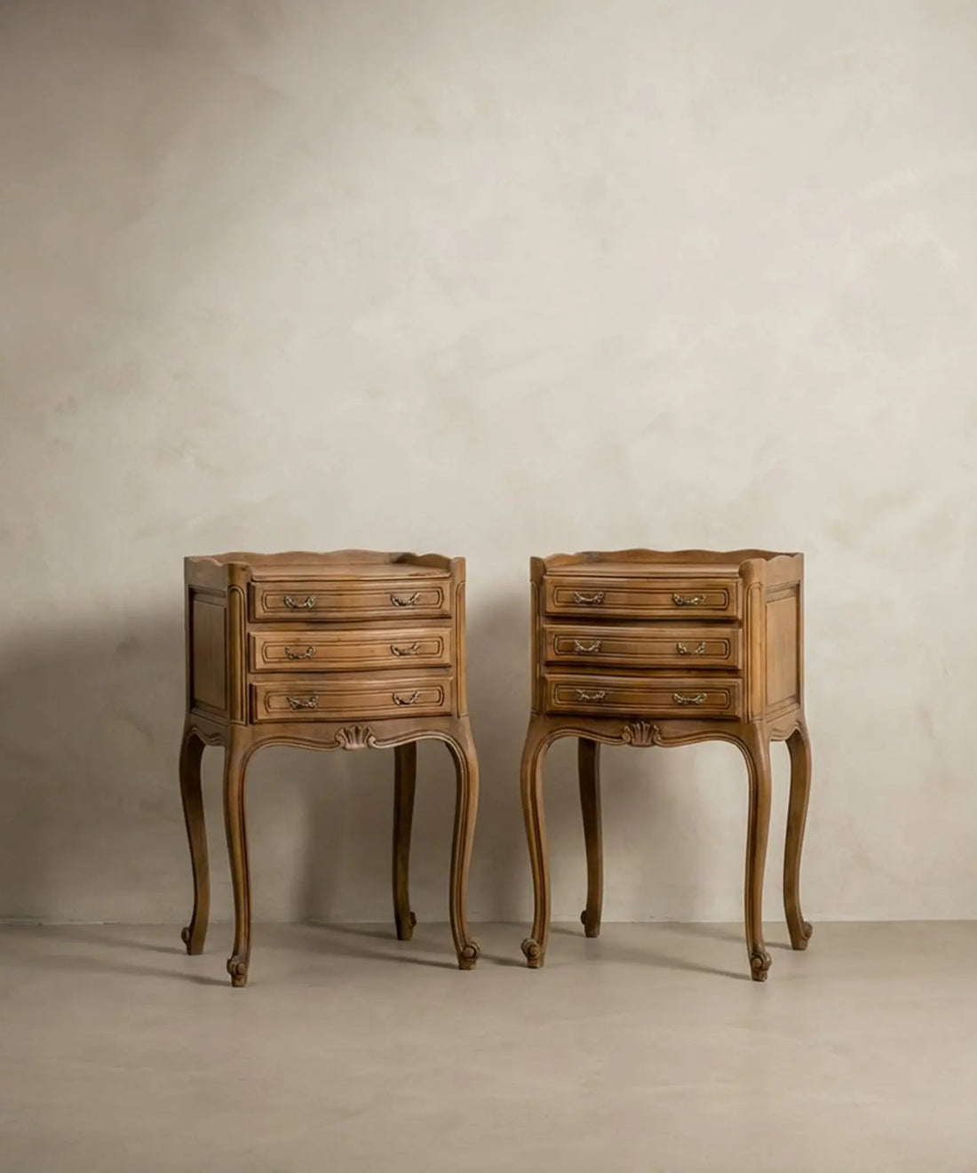 Vista frontal de pareja de mesitas de noche estilo Luis XV en madera natural clara sobre fondo neutro.