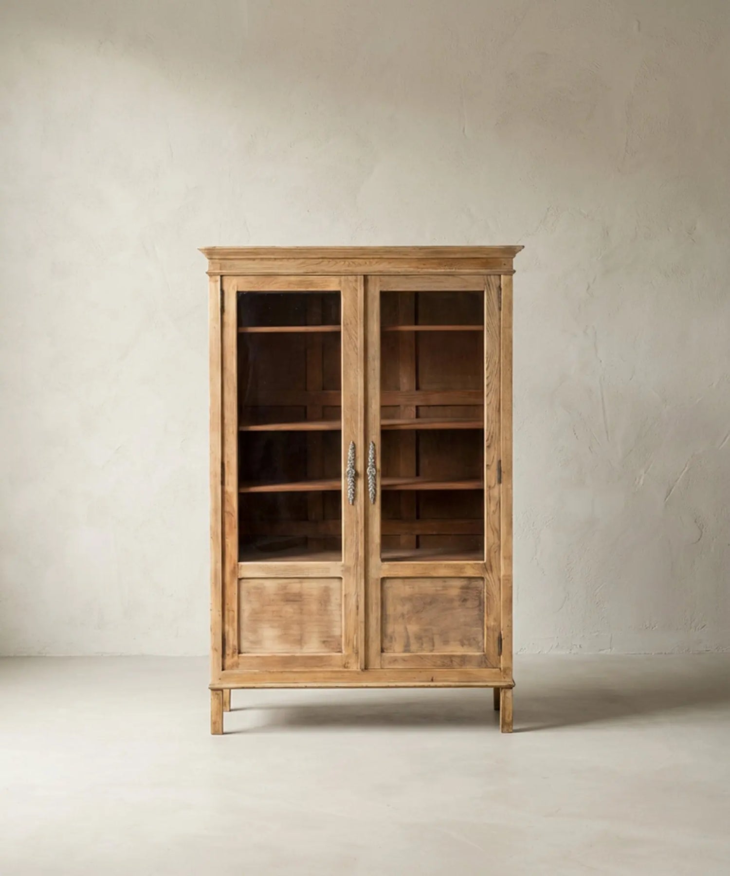 Vitrina de madera clara estilo provenzal con puertas de cristal y herrajes ornamentados sobre fondo neutro.