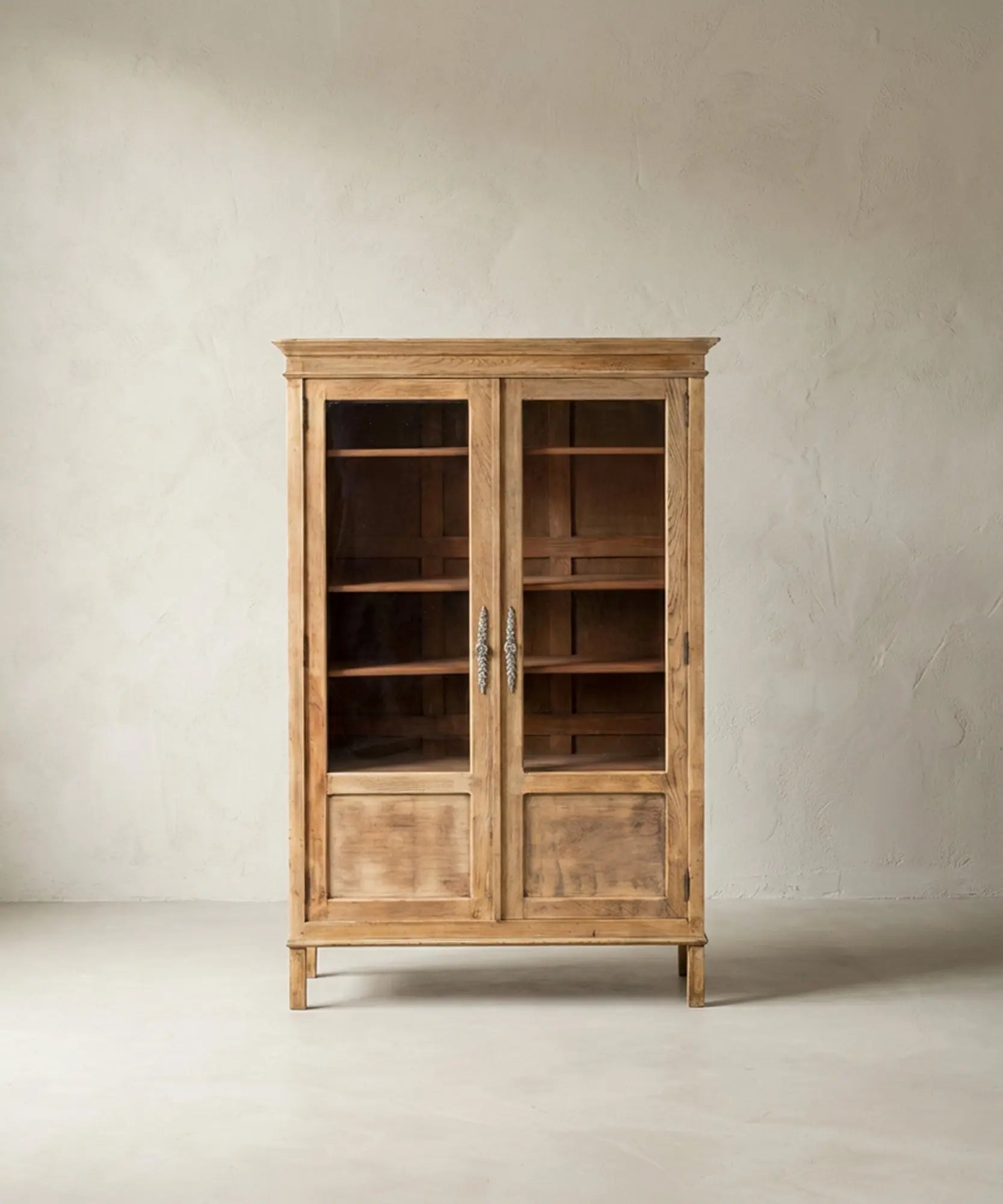 Vitrina de madera clara estilo provenzal con puertas de cristal y herrajes ornamentados sobre fondo neutro.