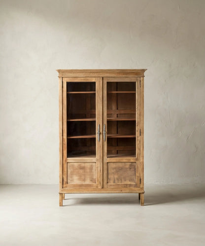 Vitrina de madera clara estilo provenzal con puertas de cristal y herrajes ornamentados sobre fondo neutro.